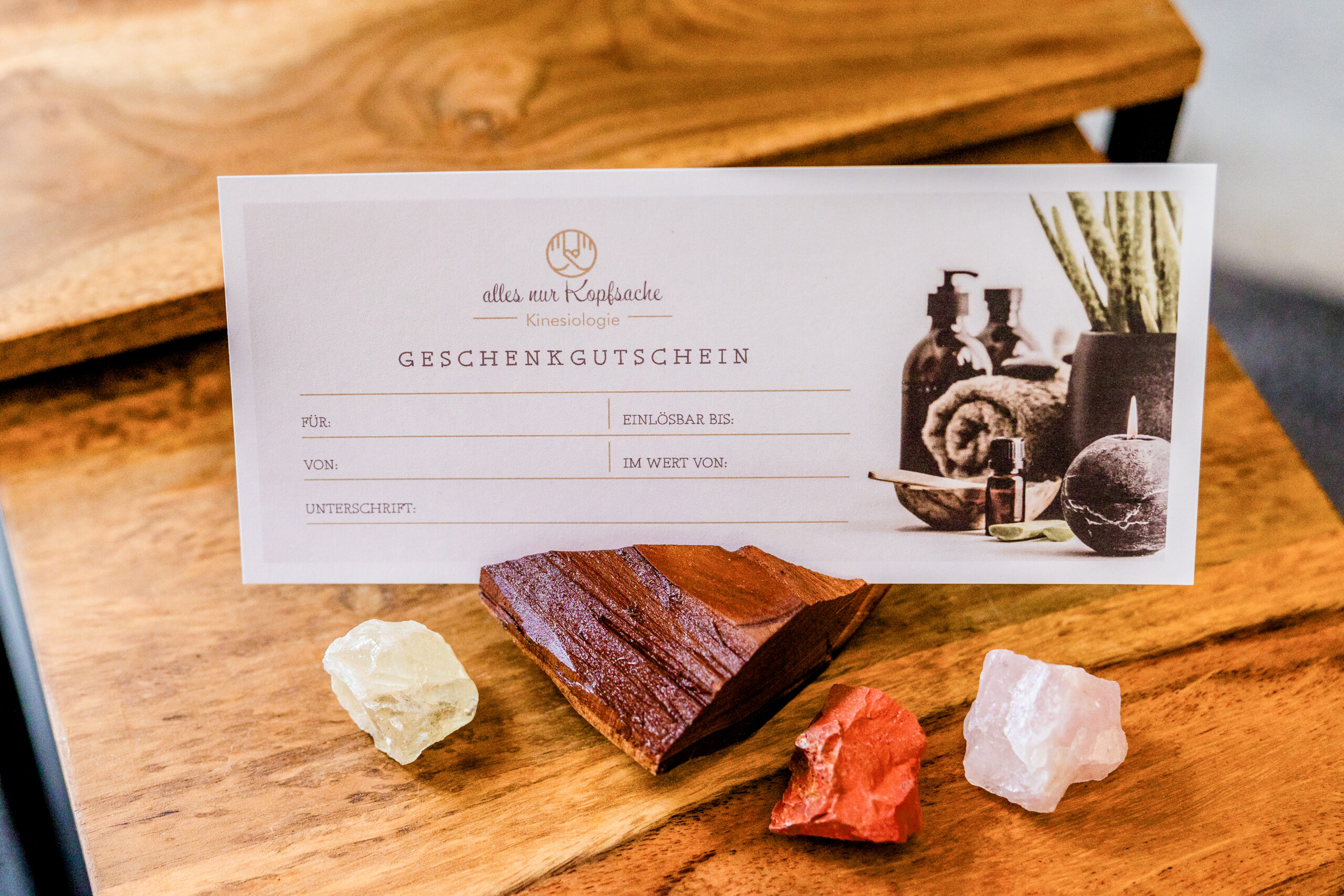 Geschenkgutschein der Praxis „Alles nur Kopfsache – Kinesiologie“ auf Holztisch mit Edelsteinen – Motiv für Kontakt/Anfrage.