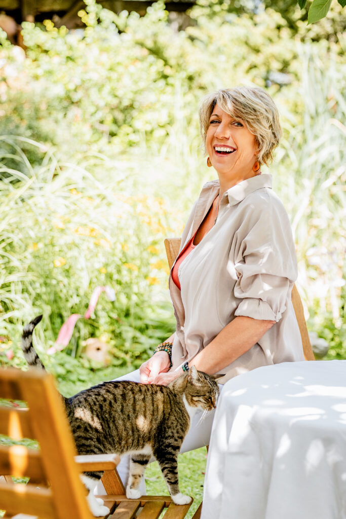 Lächelnde Therapeutin im Garten, getigerte Katze streicht am Stuhl vorbei – Über-mich-Foto der Praxis in Ebersbach.